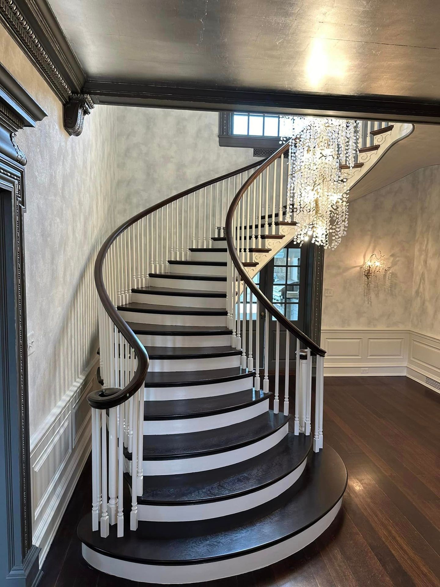 Elegant spiral staircase with dark wood treads and white railing, illuminated by a crystal chandelier.