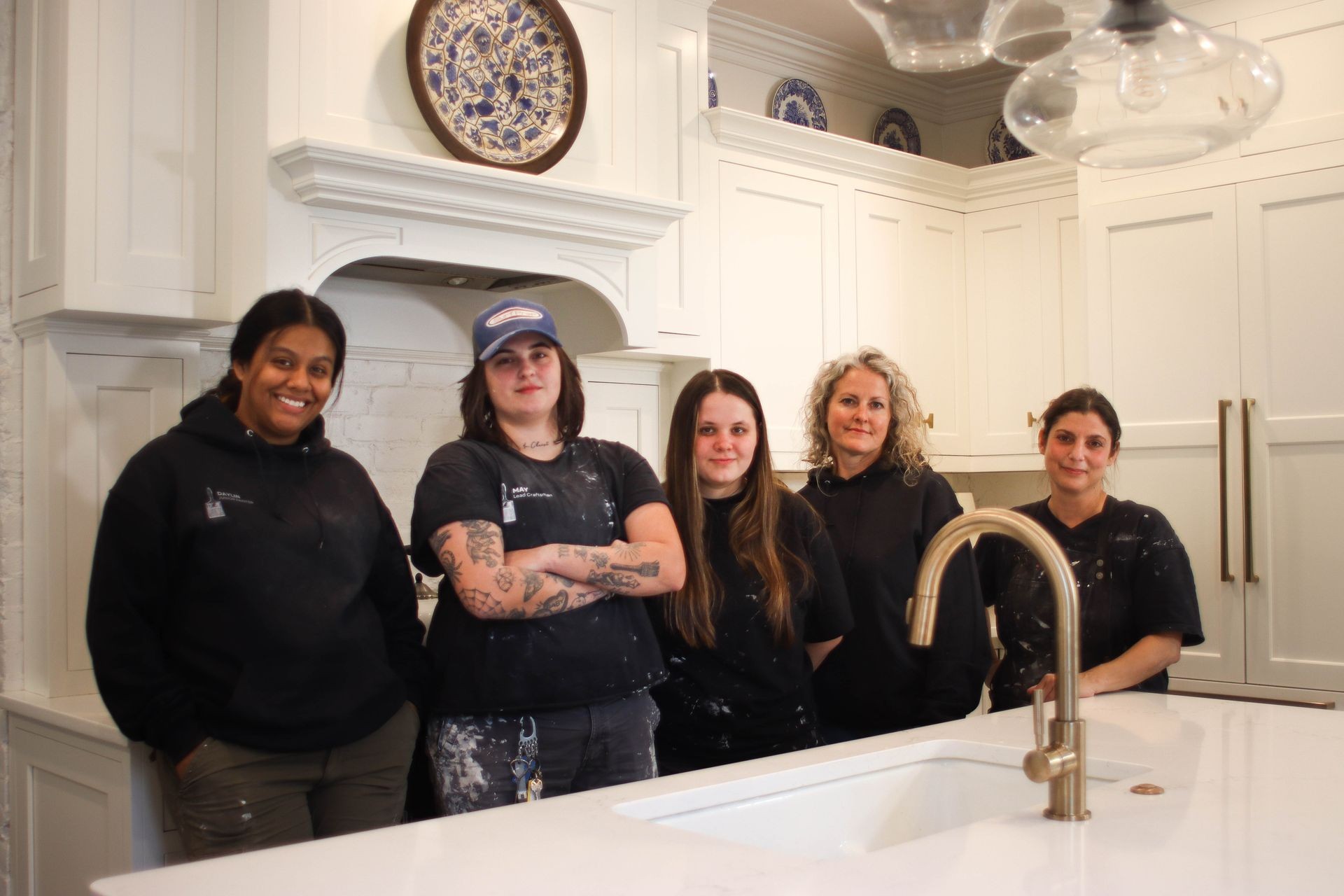 Five people standing in a kitchen with white cabinets and decorative plates displayed.